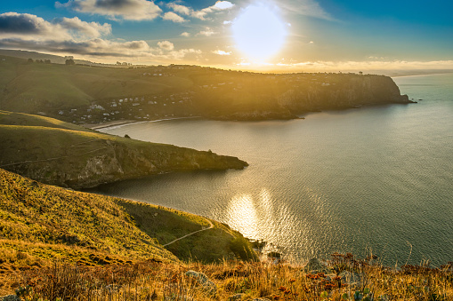 This July 2021 image was taken along the Awaroa Godley Head Loop Track, a part of Horomaka Banks Peninsula in Ōtautahi Christchurch, Aotearoa New Zealand. The setting sun reflects off Te Moana-nui-a-Kiwa Pacific Ocean. The seaside community of Te Onepoto Taylors Mistake is in the distance.