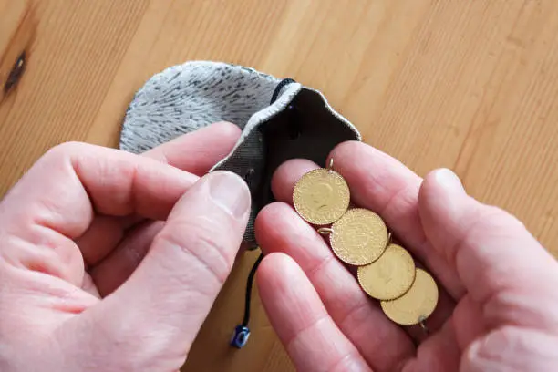 The man pours the Turkish gold coins into the pouch. Gold coins in grey bag on wooden background. Finance concept. English mean of text on coins is"Republic of Turkey Mint".
