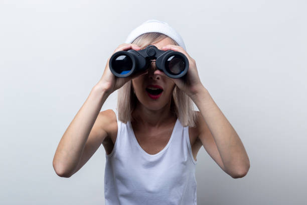 istock Surprised young woman looking through binoculars on a light background 1337031060