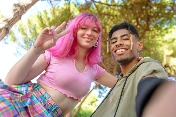 couple alternatif diversifié traînant ensemble chilling et souriant assis dans un parc posant et prenant un portrait pov selfie. amis interraciaux heureux à l’extérieur. concept d’adolescent, de joie, de plaisir et de style de vie - mode de vie alternatif photos et images de collection