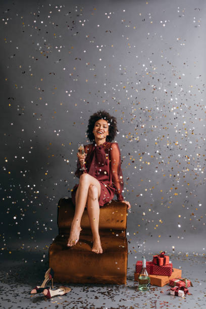 celebración de año nuevo: hermosa mujer afroamericana sentada descalza con las piernas cruzadas, sosteniendo una copa de champán bajo la lluvia de confeti - beautiful women gift christmas fotografías e imágenes de stock