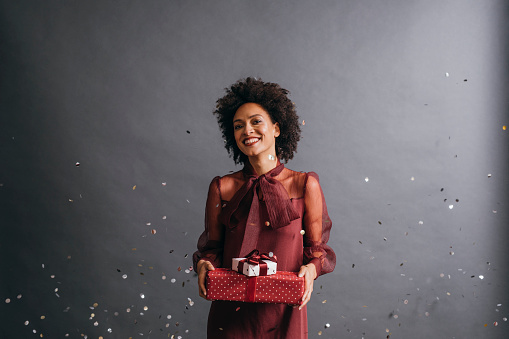 A portrait of a stunning looking African American woman holding Christmas presents in confetti rain, with a grey background