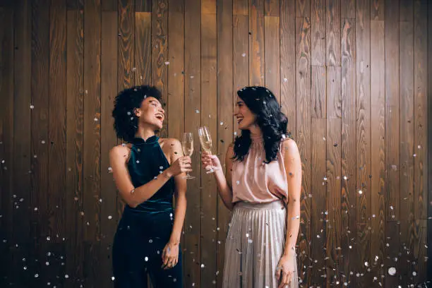 Two stunning cheerful women toasting with champagne with confetti falling all around at a party