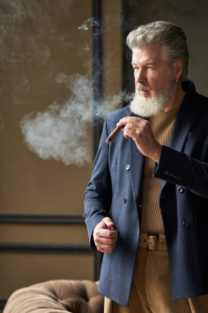 istock Stylish brutal middle aged man with beard looking away while smoking cigar, standing in loft interior 1337037404