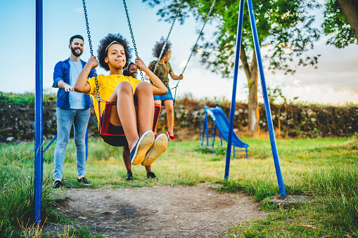 Cheerful multi-ethnic family playing at park together on swings on a summer day