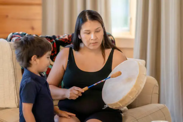 A young Indigenous mother  is playing a traditional drum for her son at home. She is sharing her family's tradition and culture with her child.
