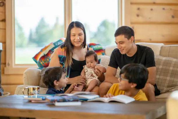 A photo of a young Indigenous Canadian family spending time together in the living room at home. The family consists of a mother, father and their three young children.