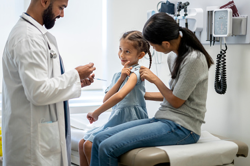 A young girl around 7 years old is at a doctor's office with her mother. They are both sitting on an examination table. She is looking at her doctor and pointing at her arm. The doctor is holding a syringe and ready to administer the flu shot onto his young patient's arm.