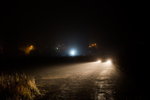 car headlight cones in night fog at field behind city building lights