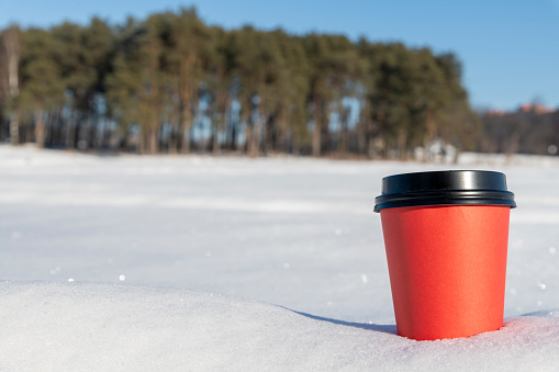 Red paper coffee cup with black lid stands outdoor on the snow. Copy space for your text and decorations. Selective focus. Blurred winter forest in the background. Take out food theme.