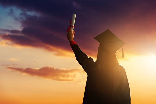 A pretty female student, celebrating her graduation and stand against the idyllic sunset view with holding her diploma high. High quality photo