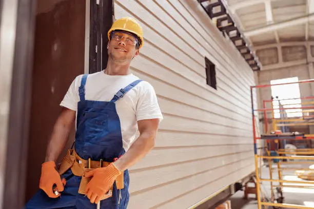 Cheerful young man construction worker looking at camera and smiling while keeping hand on tool belt