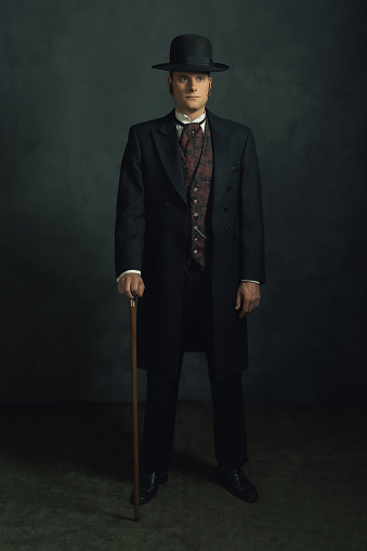 Young man in stylish vintage victorian attire. Standing with a cane in a room in front of a dark gray wall.