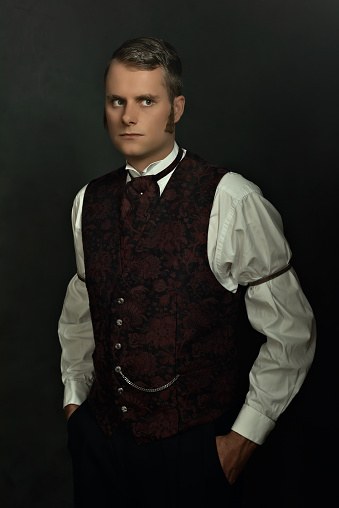 Shadowy portrait of a young man in vintage Victorian attire in front of a dark gray wall.