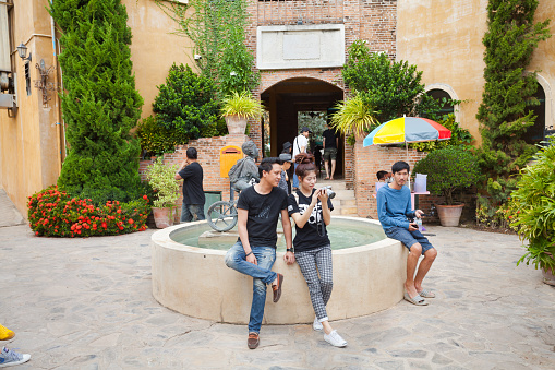 Relaxing thai couple with digital camera at fountain in Palio Khaoyai village. Couple is watching screen of camera. A man is sitting at right side. In background are peopke and a a gateway. Palio Khaoyai village is travel destination in classic italian architecture style offering pedestrian zones with stores and restaurants in valley near national park in district Mu Si, Pak Chong