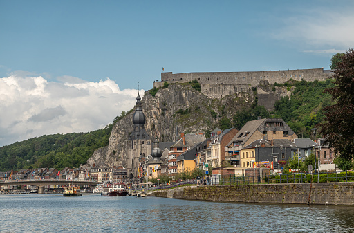 Dinant, Wallonia, Belgium - August 8, 2021: Upriver view on downtown under blue cloudscpae with dominant Citadel Fort. Boats on Meuse and green foliage around.