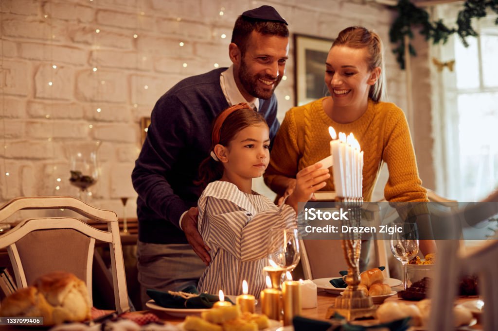 Happy Jewish family lightning the menorah before a meal at dining table. Happy parents with daughter lightning candles in menorah at dining table while celebrating Hanukkah at home. Hanukkah Stock Photo