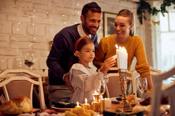 Happy parents with daughter lightning candles in menorah at dining table while celebrating Hanukkah at home.