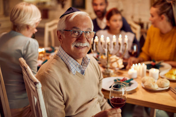 Happy Jewish senior man celebrating Hanukkah with his family at dining table. Happy senior man drinking wine while having traditional Hanukkah meal with his family at home. yarmulke photos stock pictures, royalty-free photos & images
