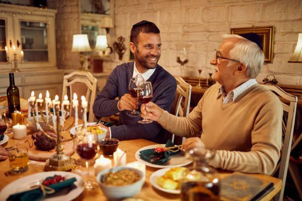 Happy Jewish men toasting with wine at dining table on Hanukkah. Happy man and his senior father toasting with kosher wine during traditional Hanukkah meal at dining table. yarmulke photos stock pictures, royalty-free photos & images