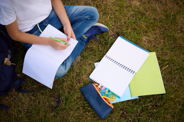 istock Top view of school child's hands writing on empty blank sheets of a workbook. Cropped image of an elementary aged child doing homework school task at the park. School supplies lying on grass 1337100596