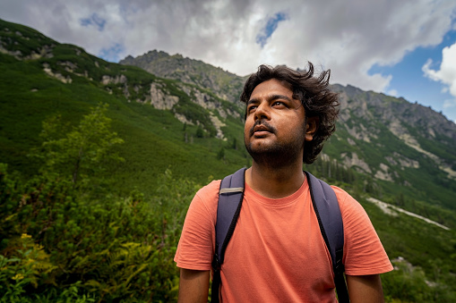 Portrait of an adventurer Indian tourist man looking up towards the way forward with bag pack against mountains during hike. Summer adventure and sports concept
