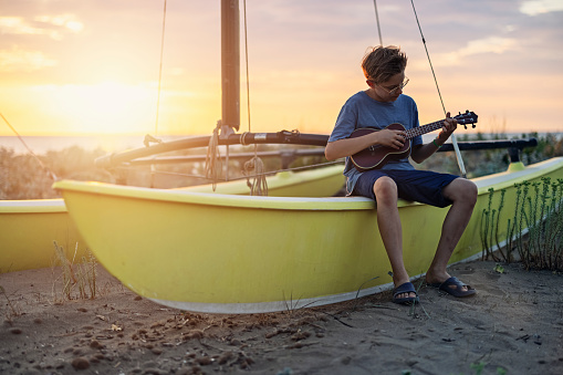 Cute teenage boy aged 11 playing ukulele. The boy is sitting on a catamaran on a beach.
Shot with Canon R5