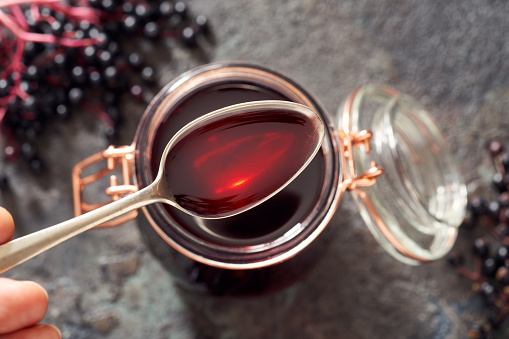 Spoon with homemade black elder syrup, with fresh elderberries in the background, top view