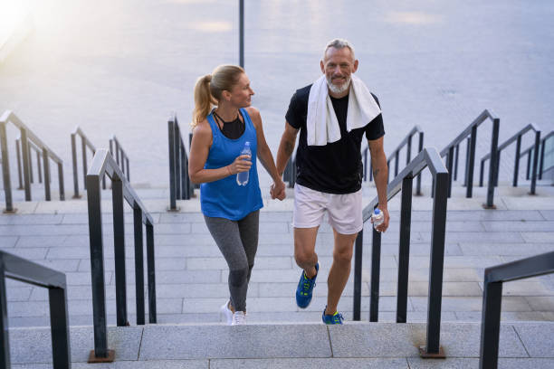 sportowa zakochana para w średnim wieku, mężczyzna i kobieta w odzieży sportowej wyglądający na szczęśliwych, trzymających się za ręce podczas wchodzenia po schodach po treningu razem na świeżym powietrzu - determination running staircase jogging zdjęcia i obrazy z banku zdjęć