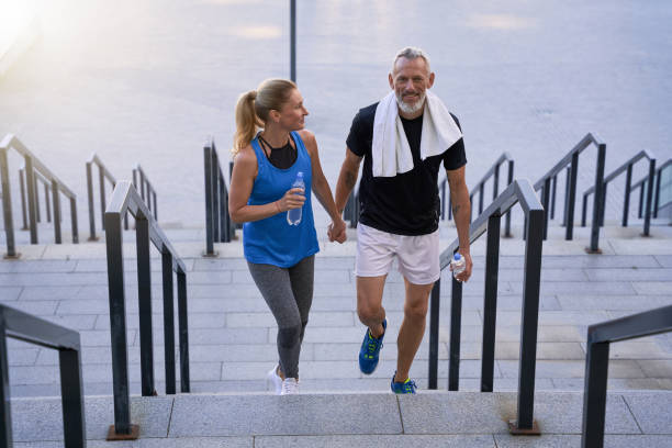 sporty middle aged couple in love, man and woman in sportswear looking happy, holding hands while walking up the stairs after training together outdoors - determination running staircase jogging imagens e fotografias de stock