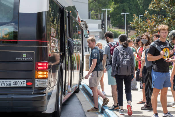 Students, some wearing face masks, board a bus at a university campus. stock photo