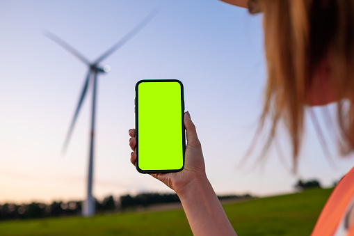 Woman engineer holding a smartphone with chromo key on the background of wind turbine at sunset