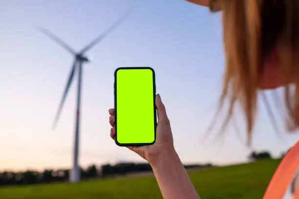 Photo of Woman engineer holding a smartphone with chromo key on the background of wind turbine at sunset.