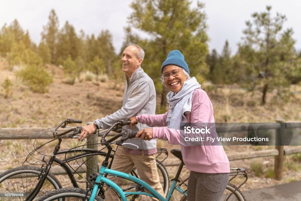 Happy And Healthy Retired Senior Couple Enjoying Vacation Together A multi ethnic senior couple are laughing together outdoors and pushing their bikes. The woman is pacific islander and the man is caucasian. Winter Stock Photo