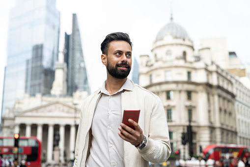 Candid portrait of bearded businessman in early 30s wearing casual summer attire, holding smart phone, and looking away from camera.