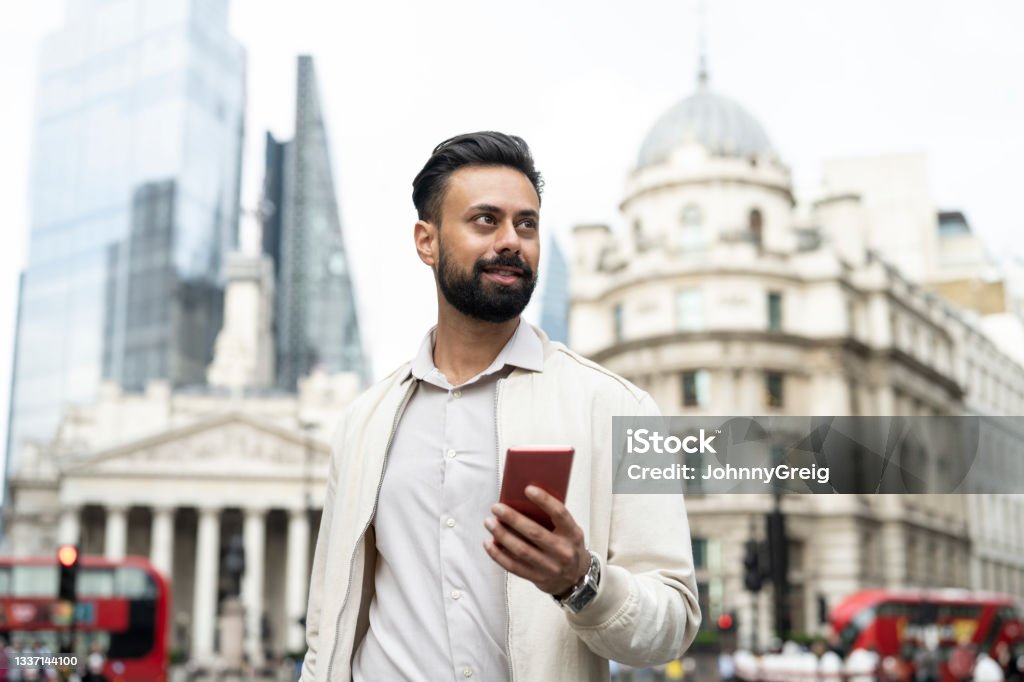 Indian man walking through London’s financial district Candid portrait of bearded businessman in early 30s wearing casual summer attire, holding smart phone, and looking away from camera. Indian Ethnicity Stock Photo