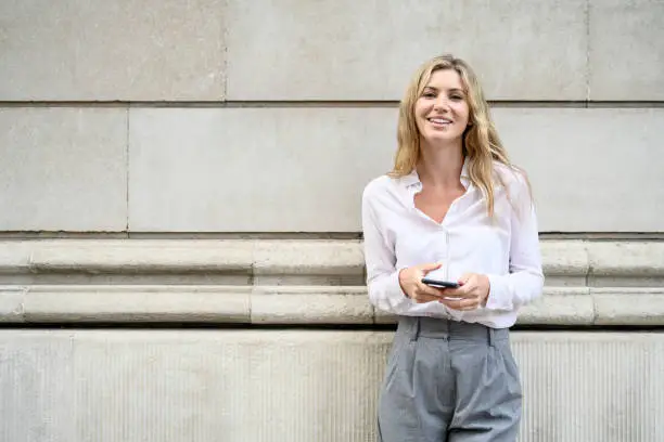 Front view of early 30s Caucasian woman in blouse and pants standing against building exterior holding mobile device, and smiling at camera.