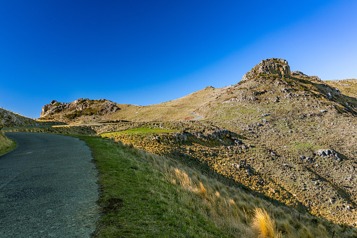 This July 2021 photo shows Summit Road as it winds through Horomaka Banks Peninsula in Ōtautahi Christchurch, Aotearoa New Zealand. Castle Rock Scenic Reserve is in the distance. This portion of Summit Road was closed to motor vehicle traffic because of damage after the 2010-2011 Canterbury Earthquakes.