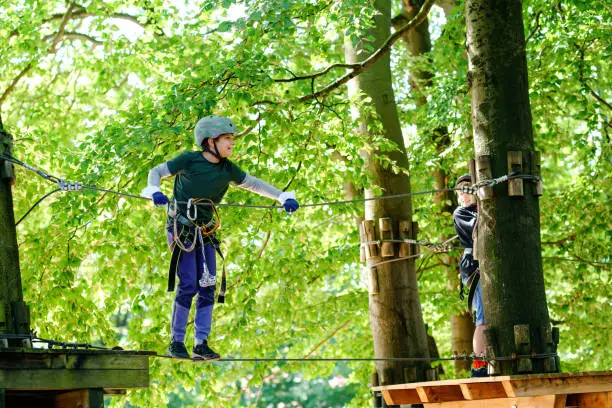 Two children in forest adventure park. Kids boys in helmet climbs on high rope trail. Agility skills and climbing outdoor amusement center for children. Outdoors activity for kid and families