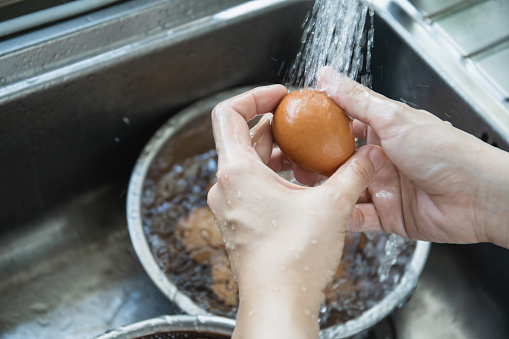 Unrecognizable Asian housewife washing a raw eggs in the sink close up, woman sanitizing and preparing a dirty eggshell before cooking in kitchen.