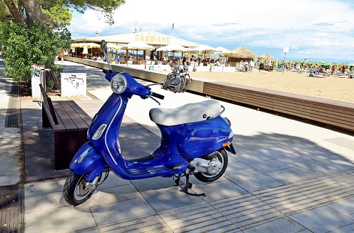 Lignano Sabbiadoro, Italy. August 29, 2021. New Vespa scooter parked outside an italian beach in summer. Beach bar Gabbiano on behind.