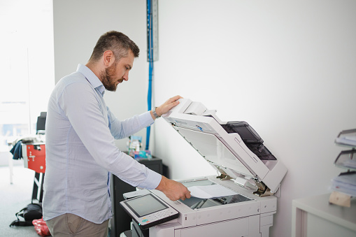 Businessman standing by office photocopier, loading the machine with documents for copying and closing lid
