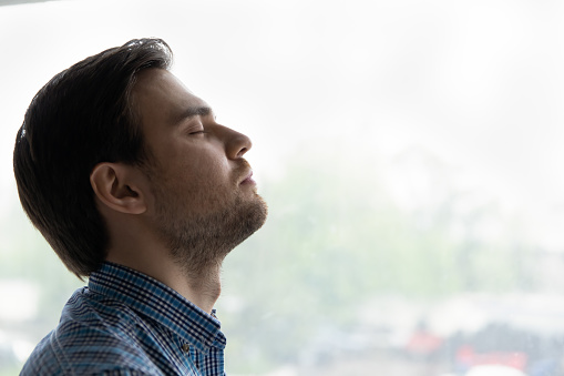 Relaxed millennial 30s man enjoying break at home, taking deep breath of fresh air with closed eyes at window, doing breathing exercises, meditation, relaxing mind, feeling stress relief. Side view