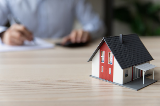Close up of small toy miniature model of house on table. Homeowner, property buyer, Real Estate Agent, mortgage broker calculating in background, counting loan, signing document, agreement. Real estate concept