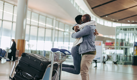 Man with face mask embracing and lifting his wife at airport terminal. African man meeting woman arriving at airport.