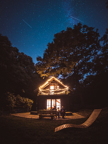 A meteor shower over the log cabin decorated with string light
Two unrecognizable people are sitting on the porch and enjoying in view
