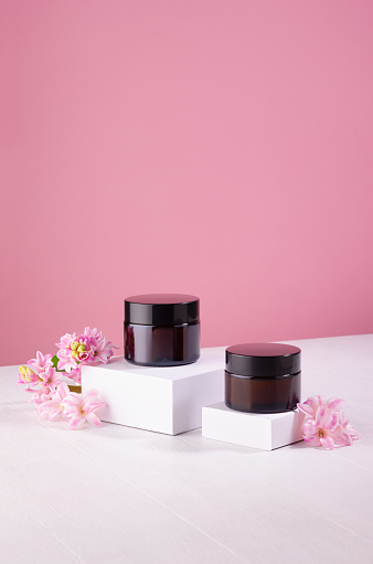 Cosmetics jars for cream of amber glass mockup on white podiums with fresh spring hyacinth flowers on pink background, vertical.
