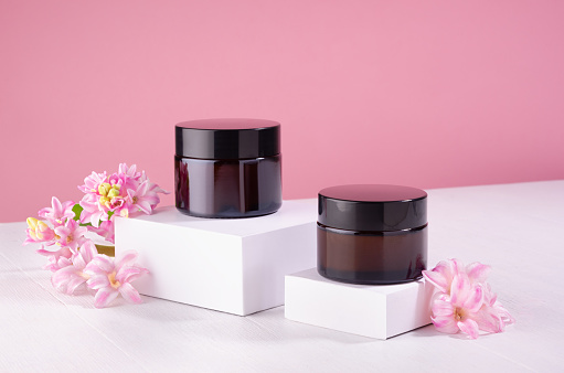 Cosmetic jars for cream of amber glass on white podium and pink wall with fresh spring flowers. Template for branding identity for cosmetics produce.