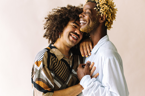 Cheerful young gay couple standing together in a studio. Two affectionate male lovers smiling cheerfully while embracing each other against a studio background. Young gay coupe being romantic.