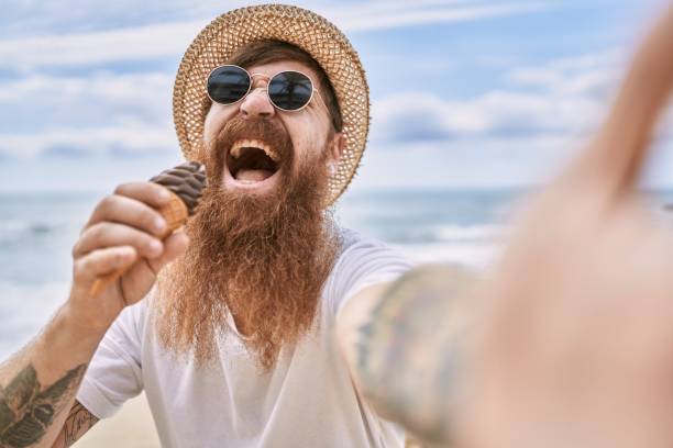 istock Young redhead man eating ice cream make selfie by the camera at the beach. 1337220811